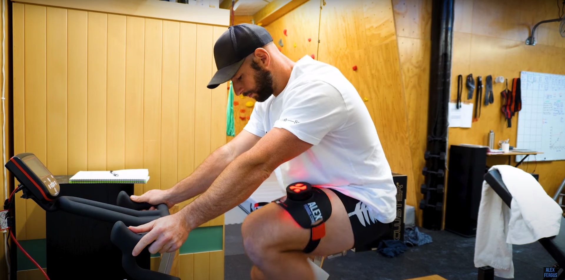 Alex Fergus using a red light therapy product on his thigh while using a stationary bike at home